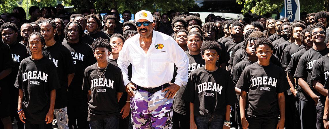 Steve Harvey stands facing the camera with a group of boys behind him